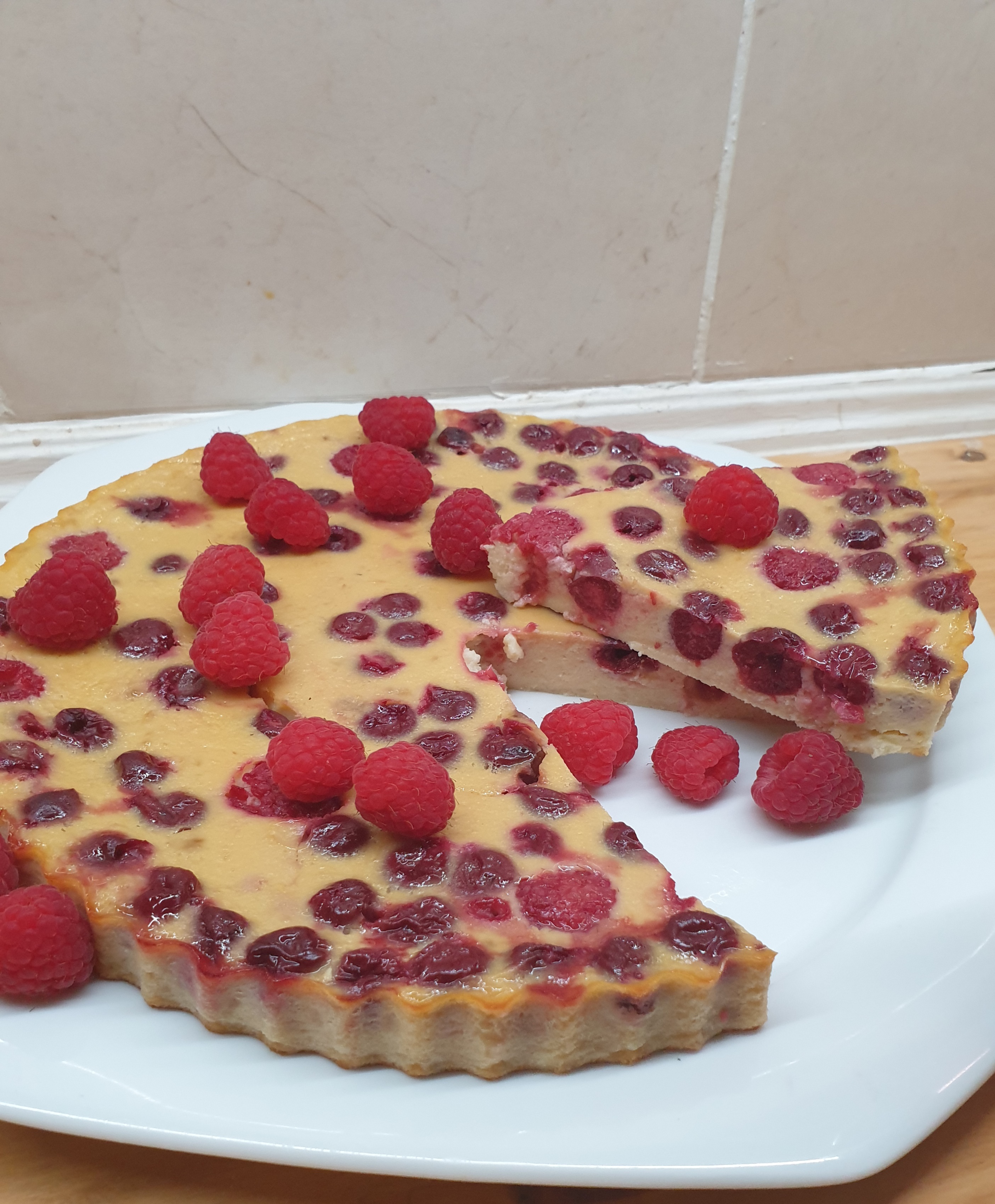 "Vers bereide flanboisier, een rijke Franse frambozen-custardtaart rijkelijk gegarneerd met verse, glanzende frambozen en een fijne laag poedersuiker, geserveerd op een wit porseleinen bord."